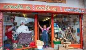 A lady stands in the doorway with her arms raised against the doorframe in the centre of the photo. There are two windows either side filled with arts and craft items.