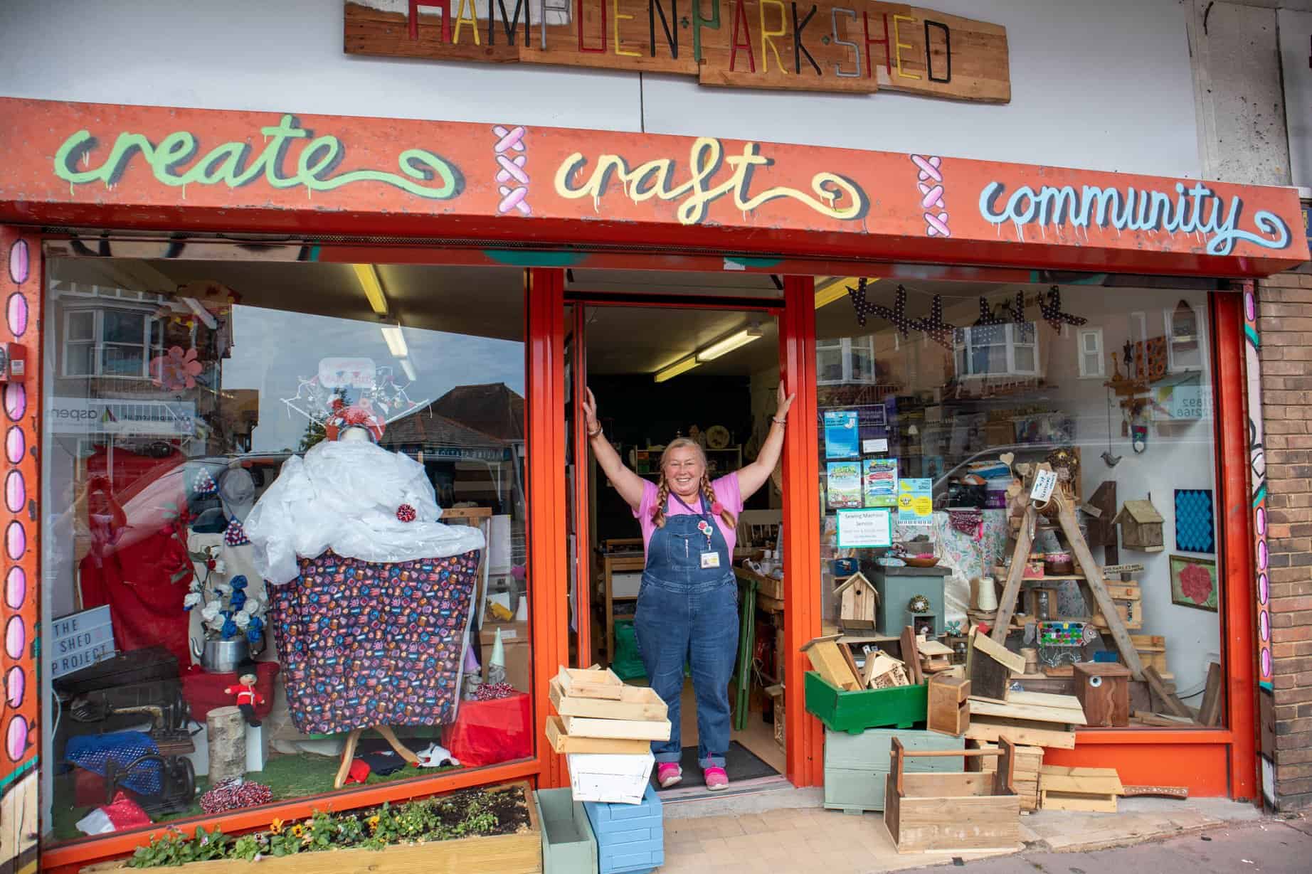 A lady stands in the doorway with her arms raised against the doorframe in the centre of the photo. There are two windows either side filled with arts and craft items.