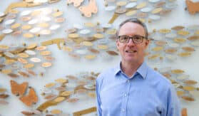 A photo of Colin, chest up, standing infront of the Memory Tree's copper, silver and gold leafs