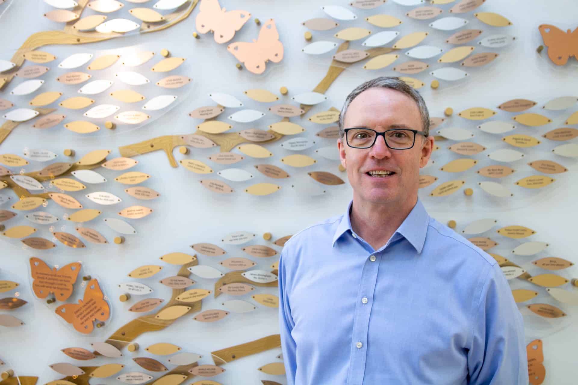 A photo of Colin, chest up, standing infront of the Memory Tree's copper, silver and gold leafs