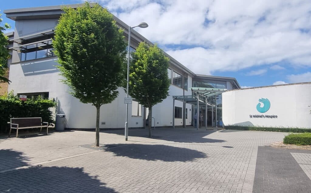 A view of the front of the hospice building, with trees lining the path, and the white building in the background.