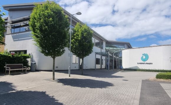 A view of the front of the hospice building, with trees lining the path, and the white building in the background.